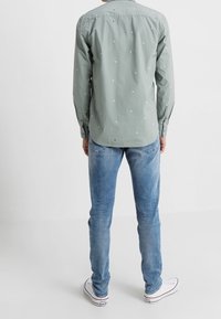 Kortärmad, ljusgrön skjorta med vita och marinblå prickar, kombinerad med medelblå jeans och vita sneakers, sedd bakifrån.