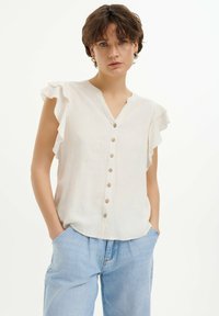 Blusa beige chiaro con maniche a volant, scollatura a V e bottoni in legno sul davanti. Sottile motivo floreale su un tessuto testurizzato.