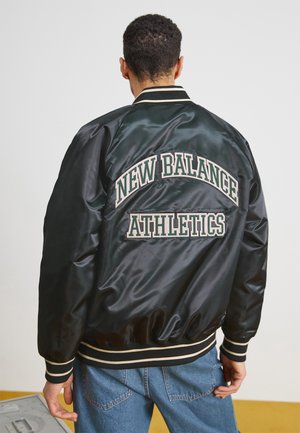 Person trägt eine schwarze Satinjacke mit dem Schriftzug "New Balance Athletics" auf dem Rücken und eine blaue Jeans, steht vor einem schlichten Hintergrund.