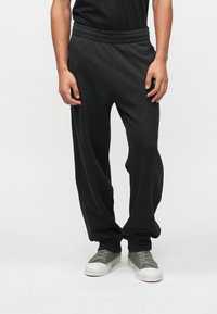 Μαύρες φαρδιές φόρμες με κορδόνι στη μέση έχουν άνετη εφαρμογή, μαλακή υφή και ελαστική ζώνη. Το μοντέλο τις συνδυάζει με γκρι αθλητικά παπούτσια για έναν χαλαρό look.