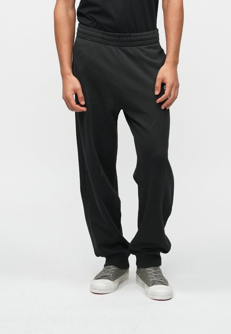 Μαύρες φαρδιές φόρμες με κορδόνι στη μέση έχουν άνετη εφαρμογή, μαλακή υφή και ελαστική ζώνη. Το μοντέλο τις συνδυάζει με γκρι αθλητικά παπούτσια για έναν χαλαρό look.