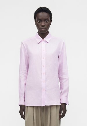 Γυναίκα που φοράει ένα ανοιχτό ροζ πουκάμισο με κουμπιά και γιακά και μπεζ πλισέ παντελόνια, στέκεται μπροστά σε λευκό φόντο.