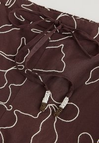 Tissu marron avec des motifs de couture abstraits blancs, bouts de cordon noués ornés de perles blanches et bronze.