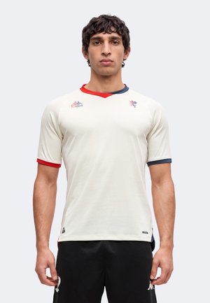 Homme aux cheveux bouclés foncés portant un maillot de sport blanc avec col et bordures de manches rouges et bleues, debout devant un fond uni.