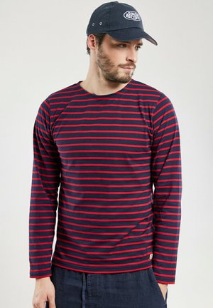 Homme portant une chemise à manches longues rayée bleu marine et rouge et une casquette bleu marine, regardant sur le côté avec une main dans la poche.