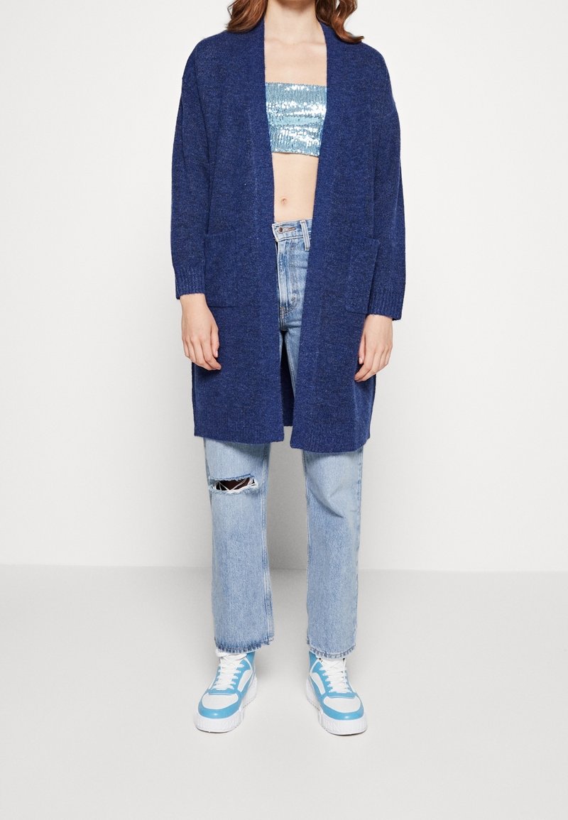 Marinblå stickad kofta med långa ärmar, sidofickor och en avslappnad passform, kombinerad med ljusblå trasiga jeans och blå sneakers.