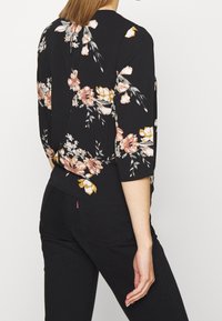 Blusa nera con motivo floreale nei colori rosa, bianco e oro. Presenta un collo rotondo, maniche a tre quarti e un orlo curvo.