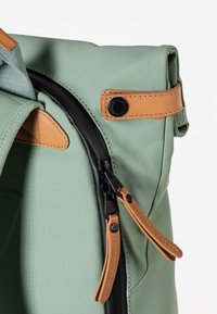 Groene canvas rugzak met tan leren accenten, zwarte rits en contrasterende kleur hardware. Bevat een zijhandgreep en leren ritslussen.