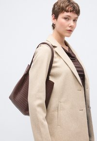 Καφέ πλεκτή δερμάτινη τσάντα ώμου με ομαλή υφή, με δύο ανθεκτικές λαβές, τοποθετημένη σε φωτεινό γκρι φόντο.