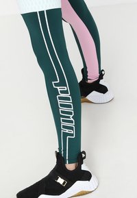 I pantaloni da allenamento con marchio PUMA presentano pannelli verde scuro e rosa con strisce e logo bianche, abbinati a scarpe sportive nere e bianche.