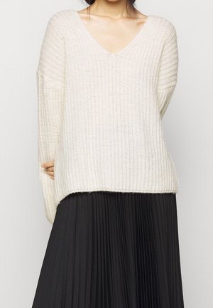 Fyldig strikket cremet sweater med V-udskæring, sænkede skuldre og ribbet tekstur, parret med en sort plisseret nederdel.