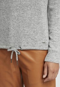 Pullover grigio a maglia con orlo regolabile e dettagli metallici, abbinato a pantaloni in pelle marrone. Il materiale sembra morbido e dalla texture interessante.