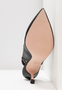Chaussure à talon aiguille noire à bout pointu, montrée par le dessous avec une semelle beige et de taille 38, positionnée debout contre un coin blanc.