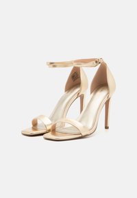 Guldmetal højhælede sandaler med to forreste stropper, firkantet tå og en ankelrem med spænde. Glat tekstur og skinnende finish.