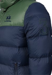 Puffjacka med gröna och marinblå färgblock, dragkedjestängning och broderad logotyp. Anmärkningsvärd design inkluderar en sydd siffra "3" på ärmen.