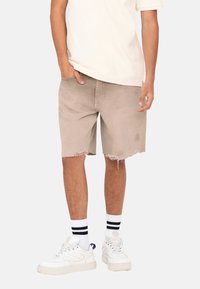 Pantalones cortos de mezclilla color beige con dobladillos deshilachados, de corte relajado, combinados con zapatillas blancas y calcetines a rayas blanco y negro. Diseño casual y hasta la rodilla.