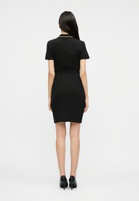 Γυναίκα στέκεται πλάτη, φορώντας ένα κοντό μαύρο φόρεμα με κοντά μανίκια και μαύρες ψηλοτάκουνες γόβες, μπροστά σε ένα απλό φόντο.