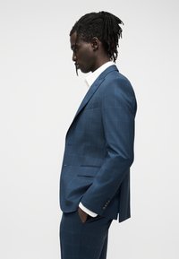 Άνδρας με ράστα, φορώντας μπλε καρό κοστούμι και λευκό πουκάμισο, στέκεται προφίλ με τα χέρια στις τσέπες μπροστά σε απλό φόντο.