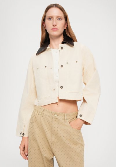 Jachetă bej cropped cu guler maro, butoni frontali și două buzunare pe piept. Purtați deasupra unei bluze albe și asortată cu pantaloni bej cu imprimeu.