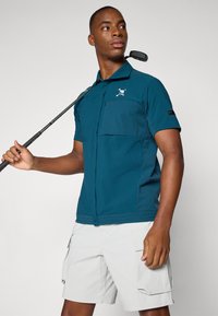 Teal short-sleeve polo shirt with a chest pocket and logo detail. Model holds a golf club, wearing light gray cargo shorts.