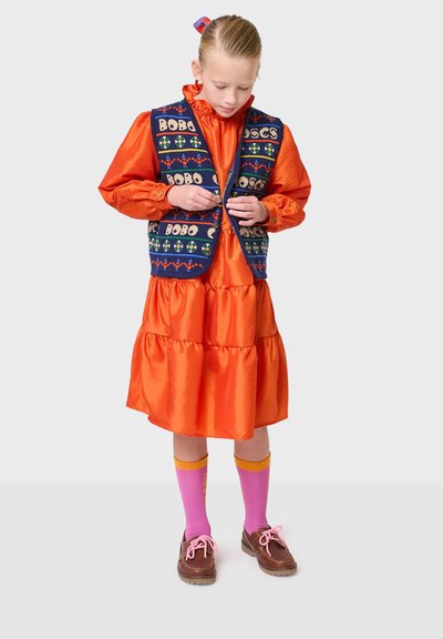 Orange satin klänning med lagerkjol, kombinerad med en marinblå stickad väst med färgglada mönster. Bruna skor och rosa strumpor med gula accenter.