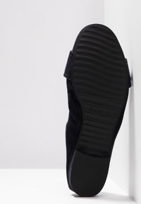 Semelle noire d'une chaussure avec motif ondulé, présentée en position verticale sur un fond blanc de mur et de sol.