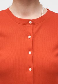 Κοντινό πλάνο ενός ατόμου που φοράει πορτοκαλί ριμπ πλεκτό τοπ με λευκά κουμπιά και μερικώς ορατό λευκό κολιέ με χάντρες.