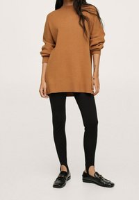 Brązowy, oversize sweter z dzianiny z opadającymi rękawami, w połączeniu z czarnymi legginsami i czarnymi mokasynami. Gładka tekstura z prążkowanymi akcentami.