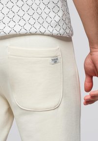 Cremfarbene Sweatpants mit einer Hintertasche und einem Etikett; glatte Textur, abgerundete Kanten und sichtbare Nahtdetails.