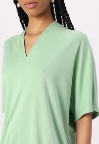 Top verde chiaro a scollo a V realizzato in tessuto morbido, con maniche corte e vestibilità rilassata. Texture liscia con dettagli di cucitura sottili.