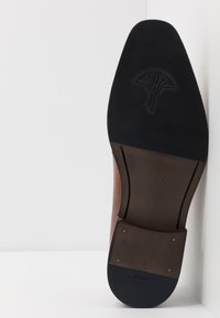 Semelle en cuir marron avec une section en caoutchouc noir texturé. Présente une empreinte de logo et des accents en métal doré. Taille indiquée comme 41.