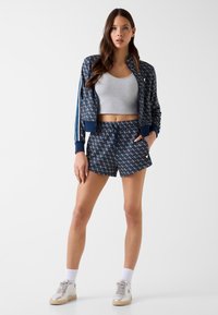Jeune femme debout, main dans la poche, portant une veste et un short à motifs bleu marine, un crop top gris, des chaussettes blanches et des baskets beiges.