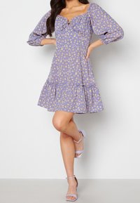 Lavendelfarbenes Kleid mit kleinem gelbem Blumenmuster, Puffärmeln, quadratischem Ausschnitt und einem gestuften Rüschensaum. Kombiniert mit lavendelfarbenen Riemchen-Absatzschuhen.