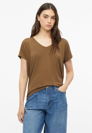 Młoda kobieta z długimi brązowymi włosami, ubrana w brązowy T-shirt z dekoltem w kształcie litery V i luźne niebieskie jeansy, stojąca z jedną ręką w kieszeni.