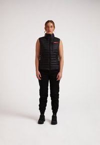 Gilet puffa smanicato nero con design trapuntato, collo alto, zip frontale e accento rosso del logo. Abbinato a pantaloni jogger neri e sneakers.