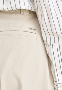 Pantaloni beige chiaro con tasca posteriore e piccolo etichetta Calvin Klein abbinati a una camicia bianca con sottili righe verticali nere.