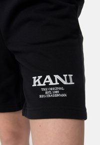Černé šortky vyrobené z bavlny, se vyšitým logem a textem "KANI" v bílé barvě. Jednoduchý design s běžným střihem a rovnými nohavicemi.