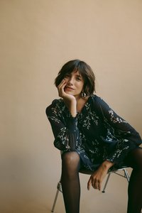 Vestito fluido con fantasia floreale scura, maniche lunghe, texture trasparente e dettagli arricciati. Modella seduta su una sedia, indossa collant neri trasparenti.