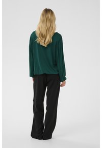 Grünes Langarmshirt mit lockerer Passform, das sanfte Falten an den Schultern aufweist, kombiniert mit schwarzen, weit geschnittenen Hosen.