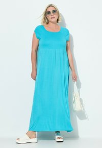 Türkises Maxikleid mit kurzen, gerafften Ärmeln und einem fließenden A-Linien-Silhouette. Kombiniert mit weißen Plateausandalen und einer weißen Handtasche.