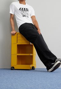 Gul förvaringsvagn med tre lådor på hjul, placerad bredvid en person som bär en vit t-shirt med grafik och svarta byxor, mot en ljusgrå vägg.