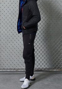 Musta tikattu takki hupulla, sinisellä vuorella, yhdistetty mustiin kapeneviin verkkarihousuihin ja valkoisiin lenkkitossuihin mustilla yksityiskohdilla. Taustalla metalliaita.