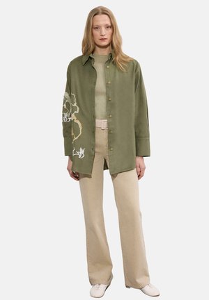 Γυναίκα που στέκεται φορώντας χακί πουκάμισο με κουμπιά και κεντήματα λουλουδιών στο μανίκι, μπεζ παντελόνι ψηλής μέσης και λευκά παπούτσια σε λευκό φόντο.