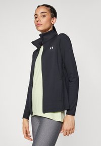 Chaqueta negra con cremallera y cuello alto, confeccionada en material suave. Presenta un pequeño logo en el pecho y mangas ajustadas, llevada sobre una camiseta ligera.