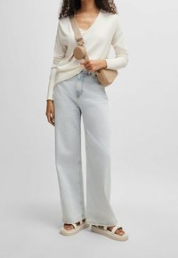 Frau trägt einen weißen V-Ausschnitt-Pullover, helle blaue weit geschnittene Jeans, eine beige Umhängetasche mit Münzfach und weiße Plateausandalen.