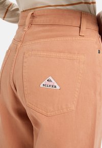 Pantalones de denim color melocotón claro con una cintura alta y corte recto. Presenta un parche triangular con la marca "QUIK SILVER" en el bolsillo trasero.