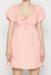 Vestido de color melocotón con mangas abullonadas, escote cuadrado, detalle de cordón en el busto y una falda relajada y acampanada. Tejido suave.