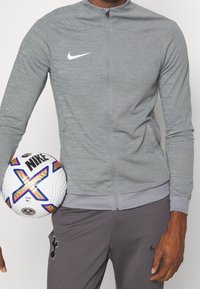 Grijze Nike jas met ritssluiting, textuurrijke stof, staande kraag en wit logo. Gehouden voetbal met meerkleurige accenten en merkembleem.