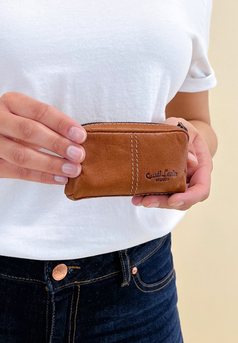 Person wearing white shirt and dark jeans holding a small brown leather zip pouch with embossed "Gusti Leder studio" logo.