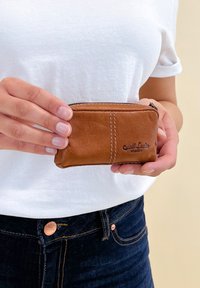 Portefeuille zippé en cuir marron avec un design cousu et un logo embossé, tenu dans une main ; texture lisse, forme compacte.